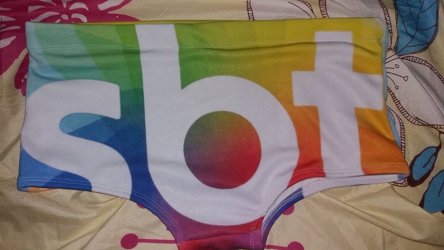 Foto colorida de uma sunga em cima de uma cama. A sunga é completamente estampada com o logo do SBT