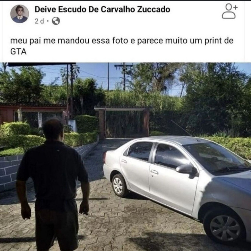 Print de post no Facebook da conta Deive Escudo de Carvalho Zuccado. Ele escreveu "meu pai me mandou essa foto e parece muito um print de GTA". A foto aparece abaixo e é uma imagem colorida de um quintal em uma casa, visto de dentro em direção ao portão, que está longe. O espaço é cheio de arbustos verdinhos e tem um carro prateado estacionado à direita da imagem. À esquerda, um homem está parado de costas para a câmera de maneira que parece um personagem de jogos como GTA, onde a câmera fica na perspectiva chamada de 3ª pessoa.